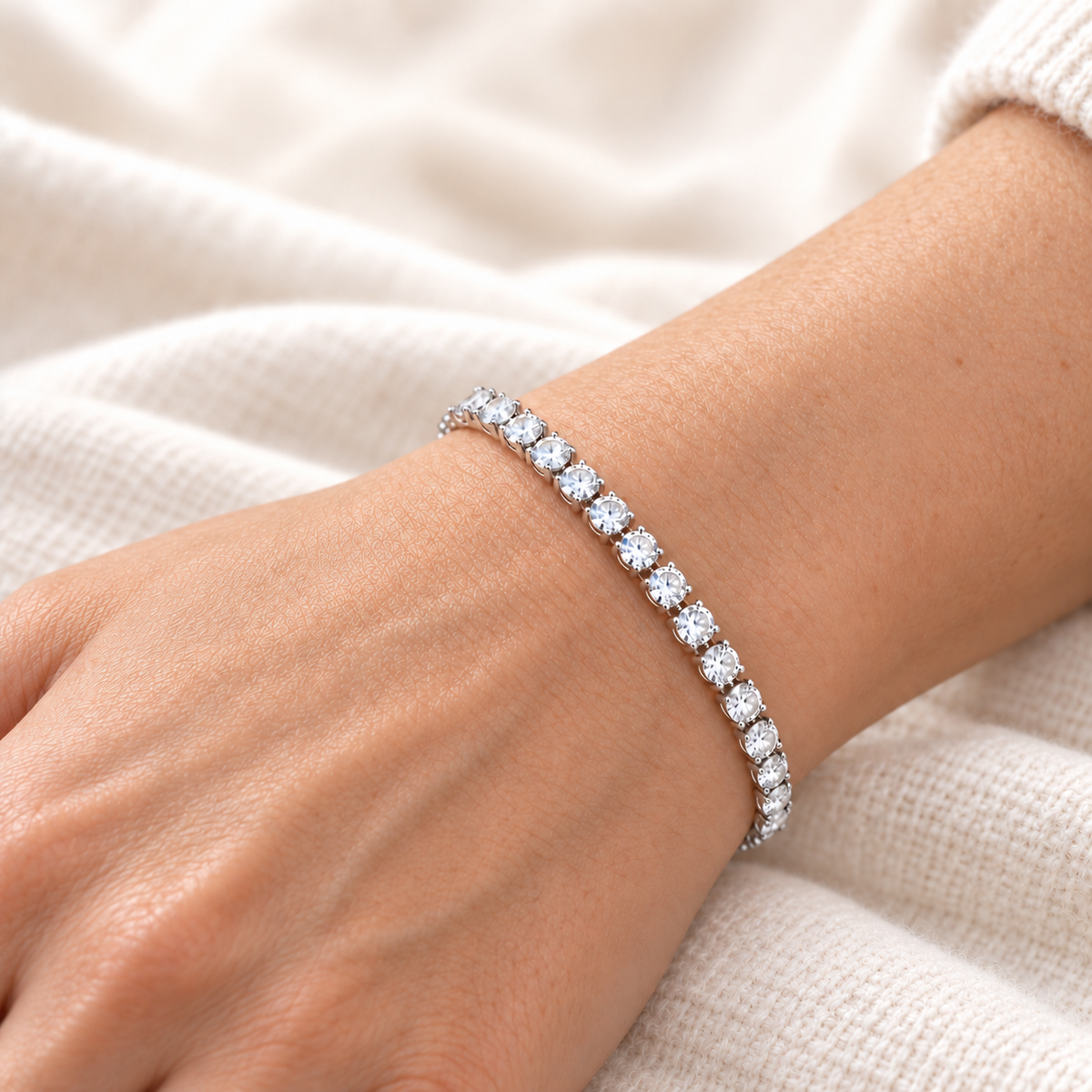 A close-up of a person's wrist wearing a silver tennis bracelet with round clear stones, set against a soft cream-colored knit fabric background.