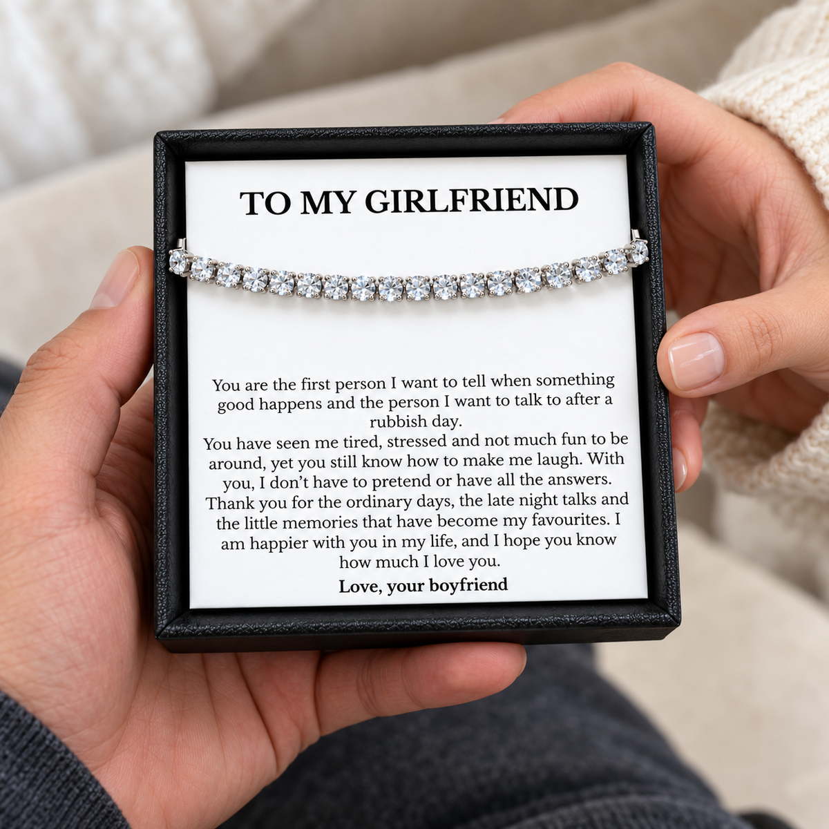 Hands holding a black gift box containing a tennis bracelet and a heartfelt To My Girlfriend message card from her boyfriend.