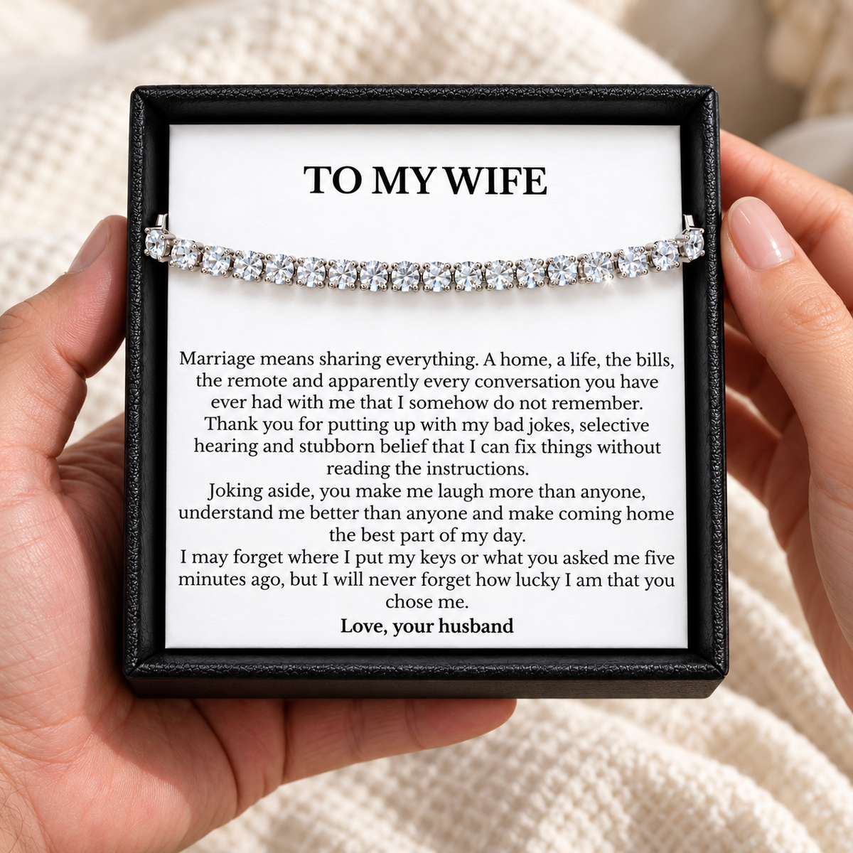 Hands holding an open black gift box containing a tennis bracelet and a humorous To My Wife message card.