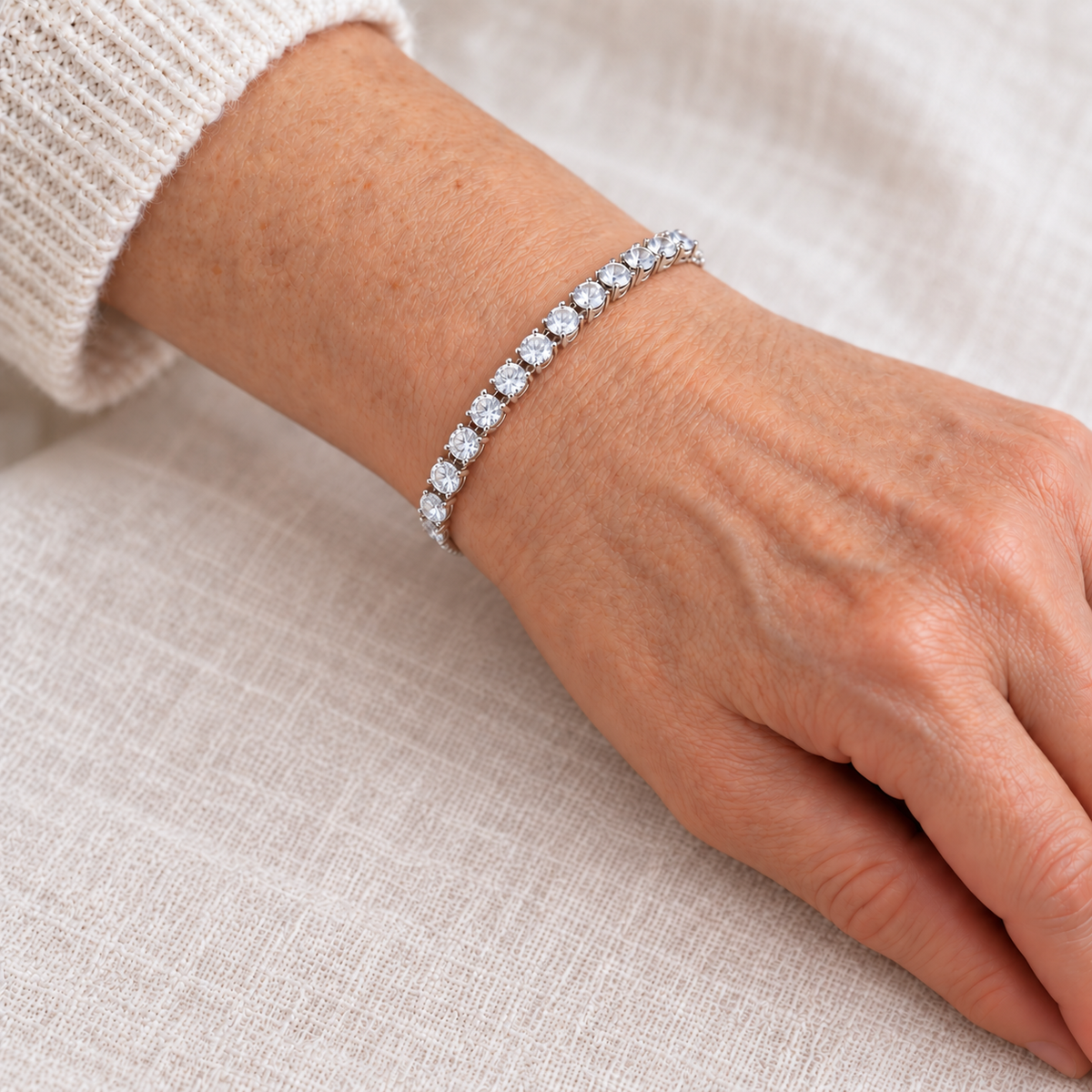 Silver-coloured tennis bracelet with a row of clear stones worn on a wrist beside a cream knitted sleeve.