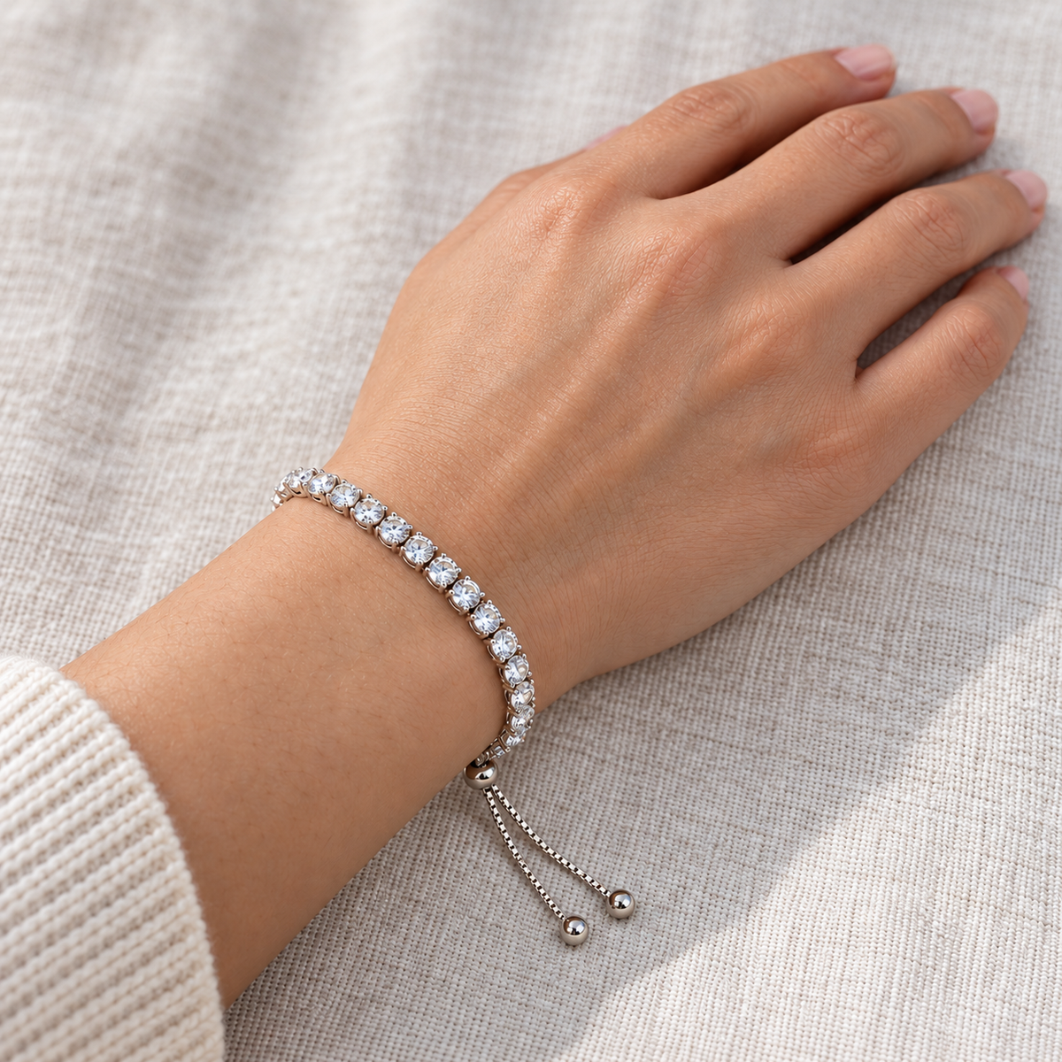 Silver-coloured tennis bracelet worn on a wrist, showing its row of clear stones, sliding adjuster and dangling chain ends.