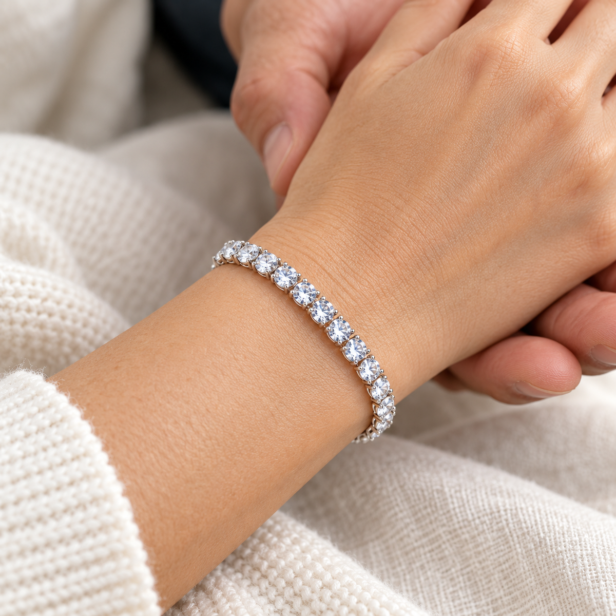 Silver-coloured tennis bracelet with a single row of clear round stones worn on a wrist while another person holds her hand.