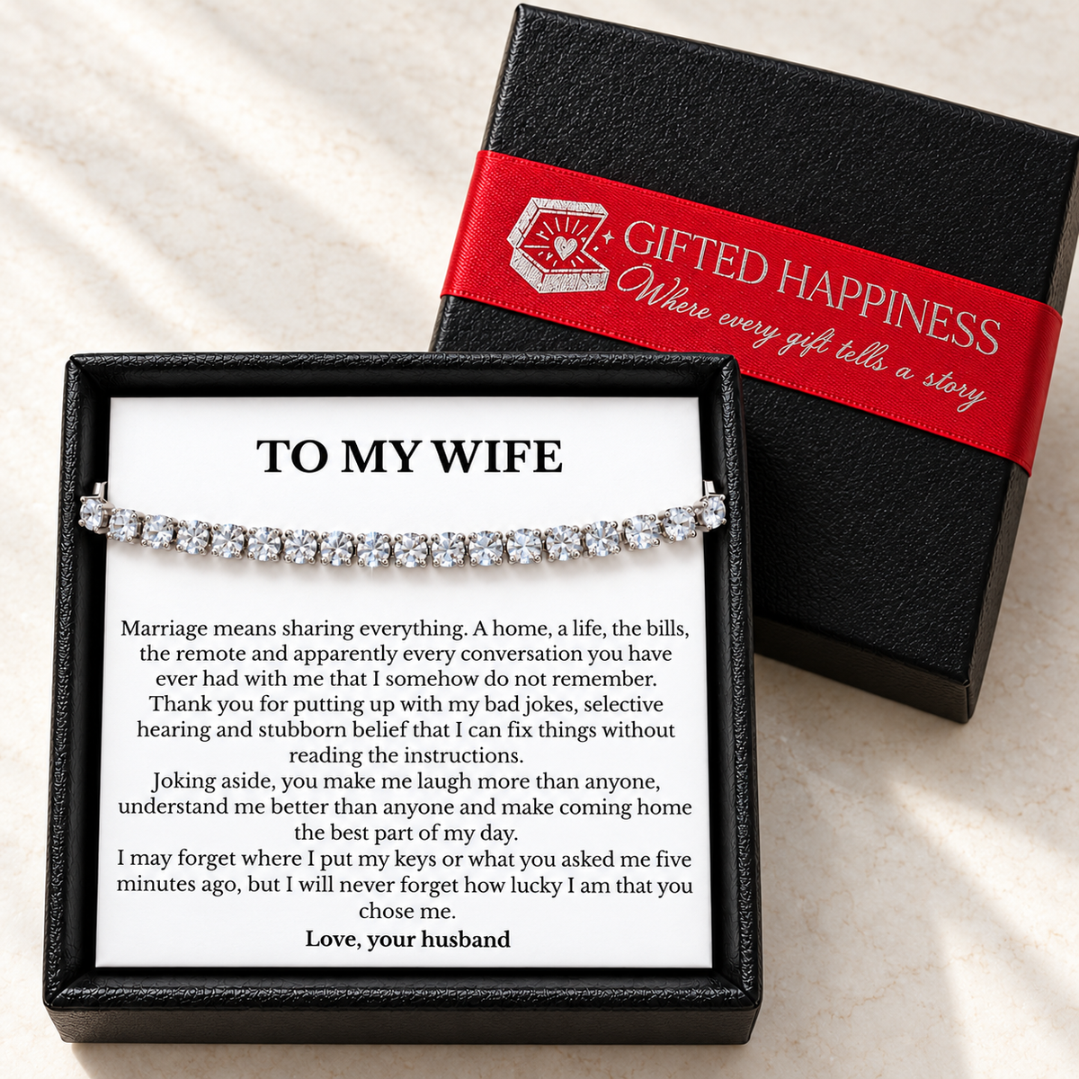 Tennis bracelet and To My Wife message card in an open black box beside its lid, wrapped with red Gifted Happiness ribbon.