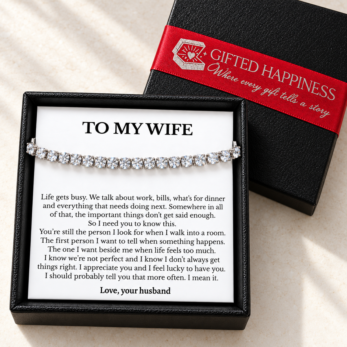 Tennis bracelet and wife message card in a black gift box beside its lid with branded red ribbon.